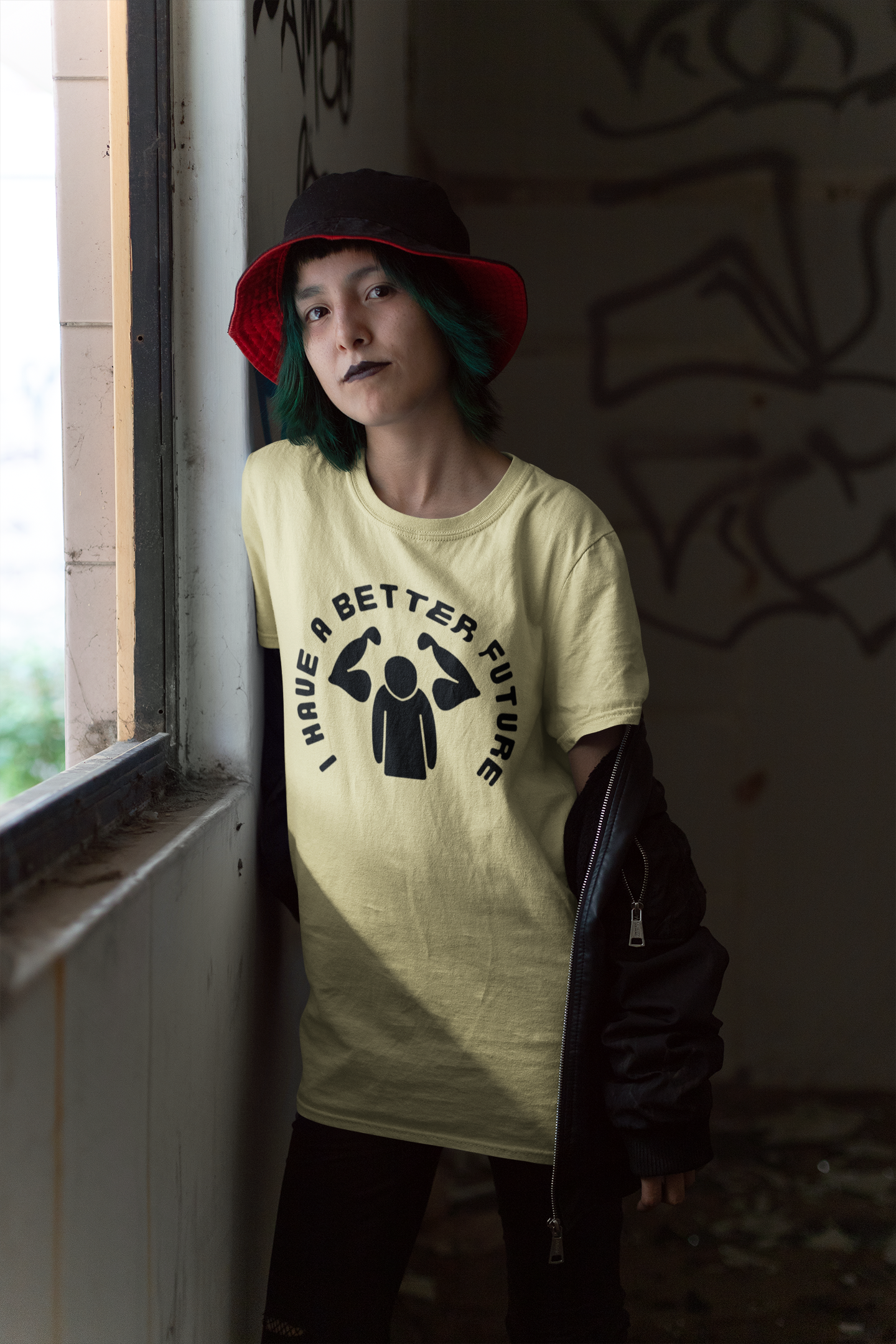 Person wearing a light-colored t-shirt with a graphic design and text, standing in a dimly lit room with graffiti on the wall.
