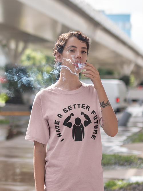 Person wearing a pink t-shirt with a graphic and text, smoking a cigarette outdoors.