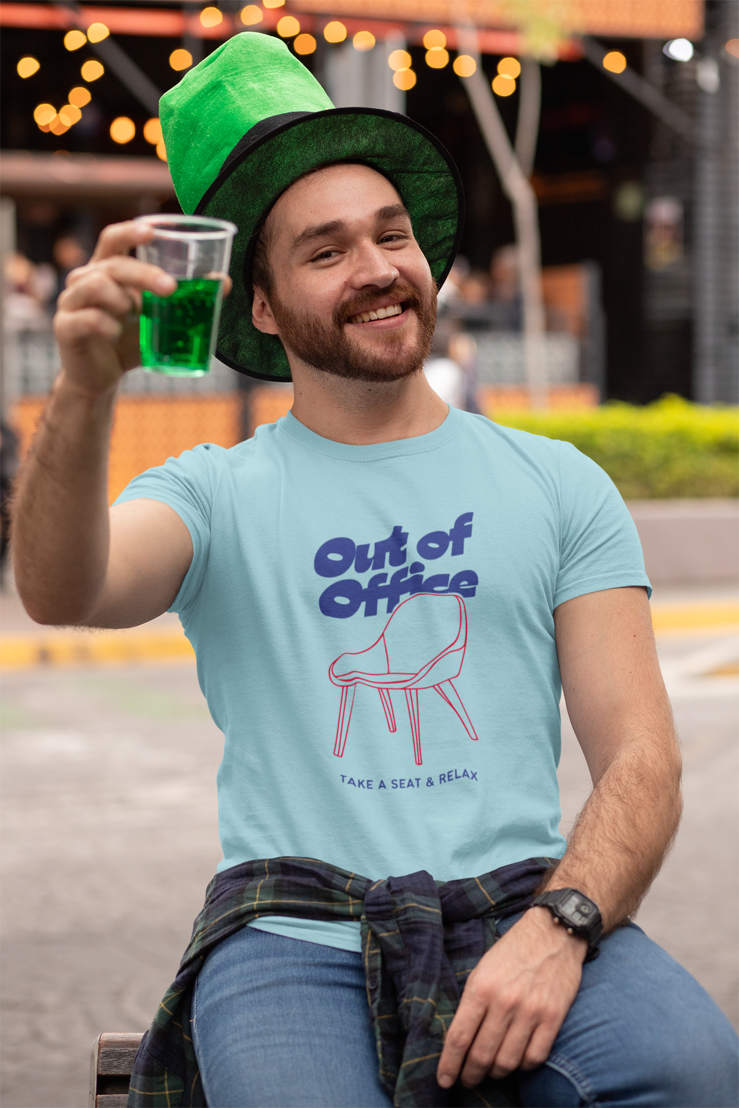 Man wearing a light blue t-shirt with a graphic and text, holding a green drink, sitting outdoors.